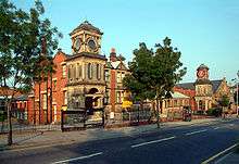 Two red-bricked buildings standing beside each other, with railings and a clock tower in front of them. The left-hand building (the school) has a sand-coloured stone tower attached to it, forming an entrance into the building. The buildings are seen from across the street, with a road and trees in front of the railings.