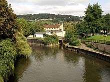 A river slowly flowing from the mid-left to bottom-right, with an entrance to a canal toward the upper right. The canal goes immediately under a small bridge, and 100 metres later under another bridge. An attractive hilled area with houses forms the background.