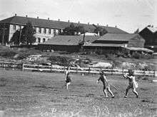 Black-and-white photo of three female baseball players. One to the right is wearing a catcher's uniform. The one in the centre is a batter who has just hit the ball. The third woman on the left is watching the two other players.