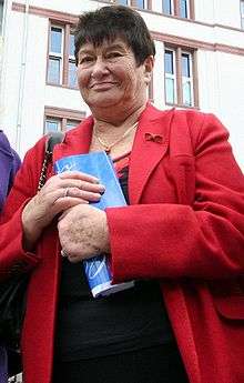 Color photograph of a older woman from her waist up. The facade of a building is in the background. The woman has short black hair. She is wearing a coat with a dress underneath, and a gold necklace with a stone pendant. She is holding a folded magazine against her chest with both hands; she has a diamond ring on the ring finger of her right hand. Her expression is a neutral one. She is somewhat overweight.