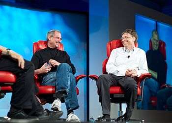 Two men in their fifties shown full length sitting in red leather chairs smiling at each other