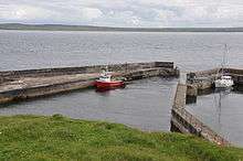 Picture of a small harbour sheltering a red and white fishing boat and a white yacht, with the mainland coast visible in the distance behind