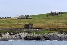 View of ruined houses and a ruined building surrounded by a stone wall, located on a green slope leading down to a rocky shoreline