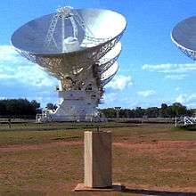 Colour photo of sandstone column with sundial atop commemorating Australian astronomer Paul Wild (foreground). Large white antenna of Australia Telescope Compact Array (radio telescope) in background; two others obscured behind it and part of another on right.