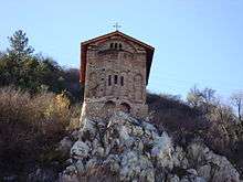 East view of a narrow medieval church topping a cliff, with some residential buildings in the foreground. The apse of the church is visible behind some rocks.