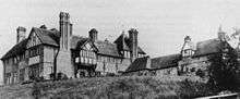 A large two-winged house seen from a low angle.  The left wing has two storeys, gables, a turret, tall chimney stacks and some timber-framing.  The right wing is lower and plainer, with a bell-cote on the end of its gable.