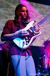 Kevin Parker playing a white electric guitar onstage, decked in purple light.