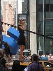 A young woman with blond, curly hair looks up while being dressed in a black cocktail dress and standing on a taxi cab.