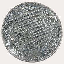 A shiny silver-white medallion with a striated surface, irregular around the outside, with a square spiral-like pattern in the middle.