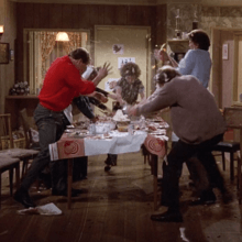 Seven people throw food pieces all over the dinner table in the dining room. The permed, brunette-haired woman in the middle owns the house.