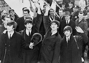 Four young men, wearing jackets and waving their hands, stand in front of an onlooking crowd.