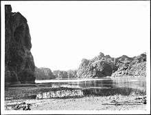 A steep rocky embankment looms in shadow at left. A tide mark is visible on the nearly vertical wall near the water line. The near bank is dried cracked mud. A few small pieces of driftwood sit on the bank. A rope lays on the bank at right. Across the placid river and in the distance rocky hills rise.