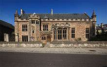 Stone building with small tower to the left of the arched doorway and bay window to the right.