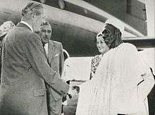 A white man in a business suit shakes hands with a black man in white robes.