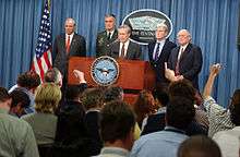 Three high-level politicians and a General, all displaying grim facial expressions, flank the main speaker.