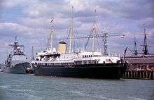 Her Majesty's Yacht Britannia is docked in Portsmouth Harbour for the 50th anniversary of the D-Day Landings in 1994. More modern Royal Navy ships are docked in behind her, and the masts of the HMS Victory can be seen in the far background.