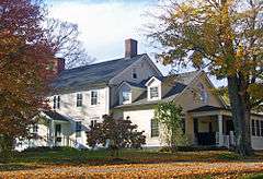 A light yellow house lit by sun from the left with trees in autumn color on either side