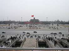 A view from above of three stone bridges leading to a sidewalk in front of a busy street. Behind the street is a large open paved area with a structure in the middle. Red flags fly from flagpoles in front