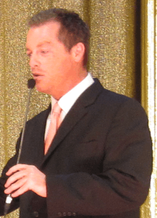 A man with dark hair wearing, including a black suite with a pink tie.