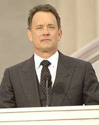 A colored photograph of a man wearing a grey blazer, with a black tie and white shirt. He is standing on a podium with a microphone positioned below his face.