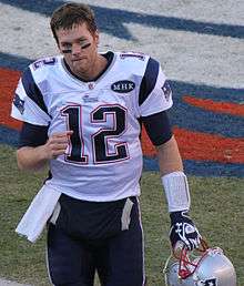 An American football player walks on a field while holding a gray helmet. He is wearing a white jersey with a blue number "12", and blue pants.