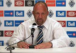 A Caucasian man with a shaved head dressed in a white shirt and striped tie sits at a table with his hands holding the base of a microphone and looks downward from the middle of the picture. Behind him are logos of various companies that sponsor the Vancouver Whitecaps.