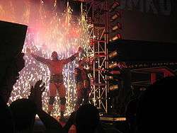 Two adult males posing on an entrance ramp at a professional wrestling event with fireworks igniting behind them.