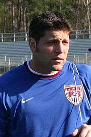 The head and torso of a man in his 30s, wearing a blue jersey. On his chest is a crest, featuring the letters "US" and a picture of a football (soccer ball).