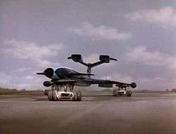 A blue, futuristic airliner rests on top of three orange support vehicles (one underneath the fuselage and two underneath the wings) as it speeds out of control along a runway against a grassy backdrop, the sky bright with clouds.