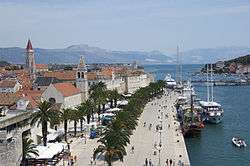 Coastal town with a palm tree promenade and houses with red roofs.