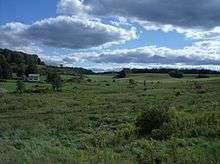 Rolling countryside with fields, some cultivated, others not. There is a small house at the left center.