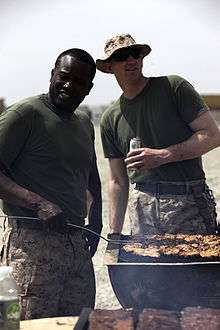 Two men in identical short-sleeved shirts and camouflage pants, one very dark-skinned with no hat and one very light-skinned wearing a hat and sunglasses, stand smiling over a barbecue full of cooking meat in a bright location.