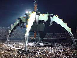 The tour stage. The video screen was above the band in a large black container. Four silver legs supporting the screen curved down into the audience. Five circular orange lights were dotted along the top of each leg. The round stage was surrounded by a semi-circular catwalk which can be reached by crossing a bridge. The audience surrounded the band on all sides.