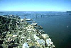 A long bridge crosses a huge river flowing into a vast body of water under a blue sky. The bridge begins in a settlement with streets, buildings, and wharfs along the river and extends out of sight toward a low hill on the far shore. The first part of the bridge has a superstructure and is high above the water but then the bridge gradually descends and continues out of sight not so far above the water.