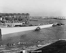 Black and white photo of a white ship on a body of water with several smaller ships near it. Industrial buildings are visible behind the ships.