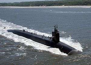 Submarine underway with a beach as a backdrop. Several people are standing atop of the boat.