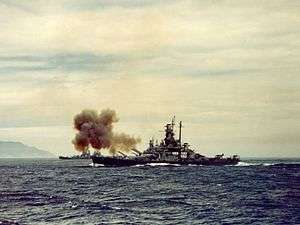 Color photo of a warship at sea. Smoke is rising from the bow of the ship, and land is visible in the background.