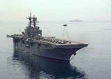 A jet aircraft hovering above flight deck of a large military ship, with several aircraft visible on the deck.