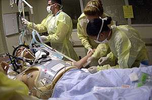 Health care providers attending to a person on a stretcher with a gunshot wound to the head. The patient is intubated, and a mechanical ventilator is visiblein the background.