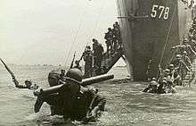 Men wearing military uniforms and carrying equipment walking down ramps from a ship into the sea