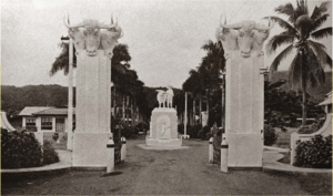 Two white pillars are topped with sculptures of bull's heads. A statue of a man holding a domestic animal can be seen beyond the pillars.