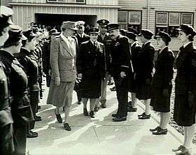 Two women and three men flanked by two lines of women in dark military uniforms