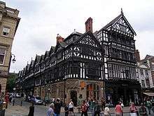 The corner of a street with a range of shops running up the right side of the street; the shops have stone lower storeys and highly detailed timber-framed upper storeys that include oriel windows and gables.  To the right of the range of shops is another shop with a modern shop front; it is taller than the others, and is timber-framed above the ground floor.