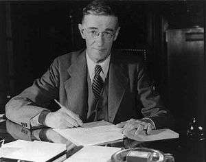 A man in a suit poses sitting at a desk, pen in hand. Papers and an ash tray are on the desk.