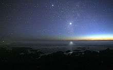 A photograph of the night sky taken from the seashore. A glimmer of sunlight is on the horizon. There are many stars visible. Venus is at the centre, much brighter than any of the stars, and its light can be seen reflected in the ocean.