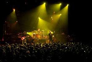 Three white Englishmen perform on a darkened stage, with yellow lights shining on them, whilst a crowd watches them.