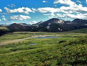 A road curving gently across a grassy landscape with a parking area along it in the center, seen from a slope above it. In the background are snow-capped peaks and a blue sky filled with fluffy clouds.