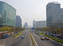 A wide divided street, seen from above, flanked by trees and medium-height high rises. The one on the left is somewhat curvilinear