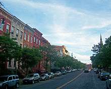 A street with three-story brick rowhouses in a variety of colors and short trees on the left, with taller trees and a distant steeple on the right.