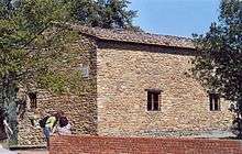 Photo of a building of rough stone with small windows, surrounded by olive trees